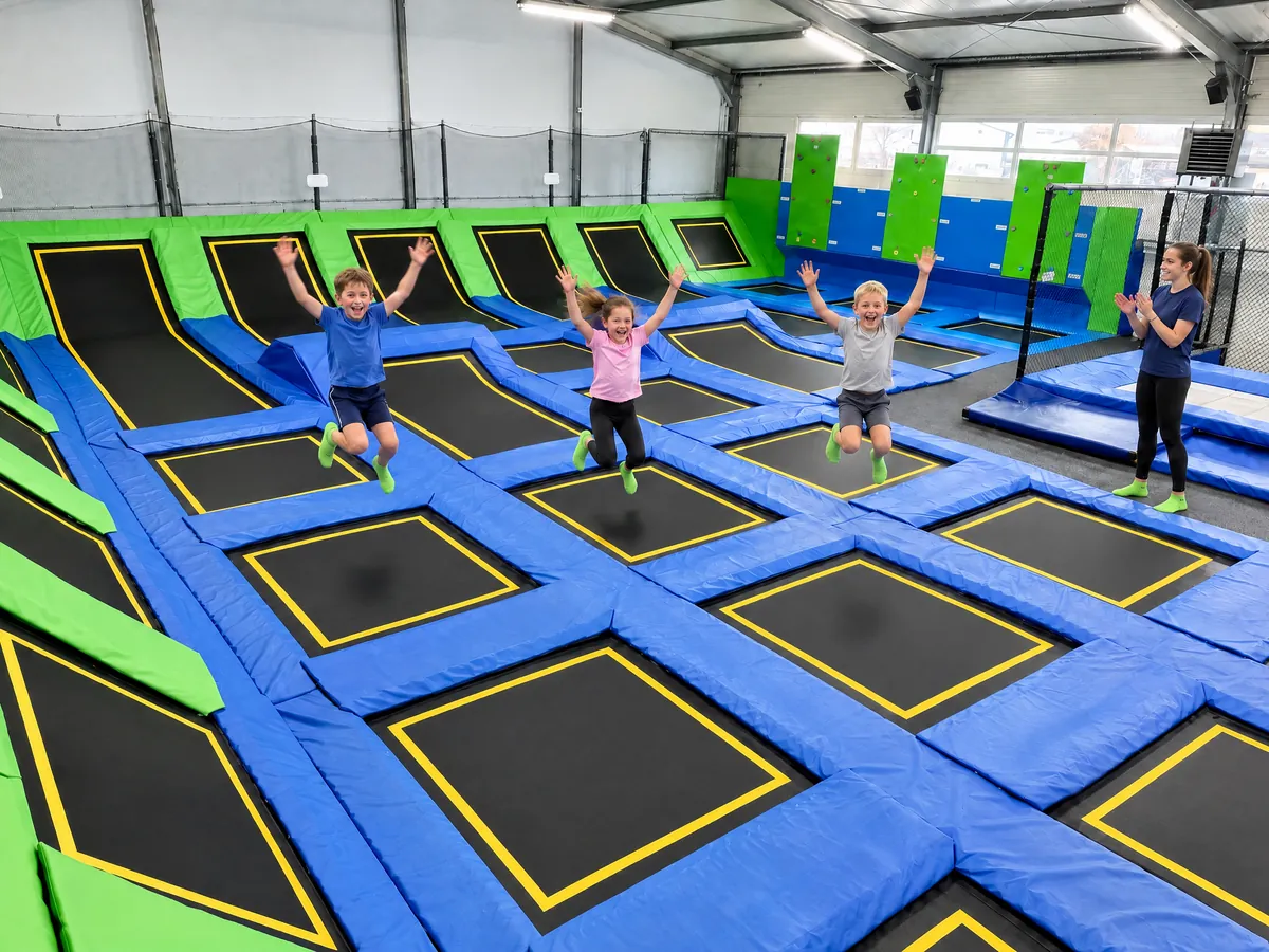 Troje dzieci w szczycie skoku nad polami trampolin, z boku trenerka bije brawo (obraz wygenerowany na tle prawdziwej hali)