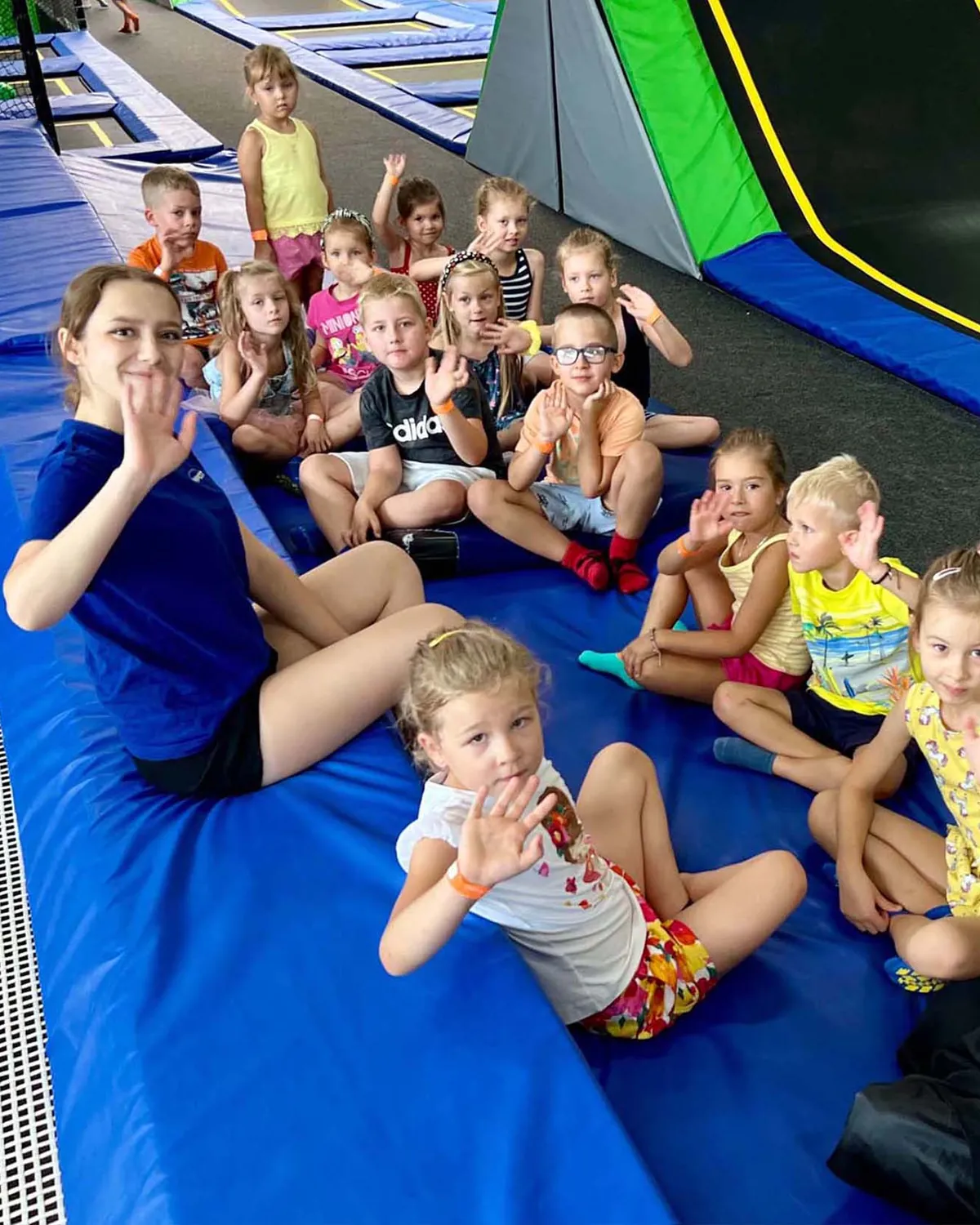 Animatorka z grupą dzieci siedzi na macie przy trampolinach, wszyscy machają do aparatu